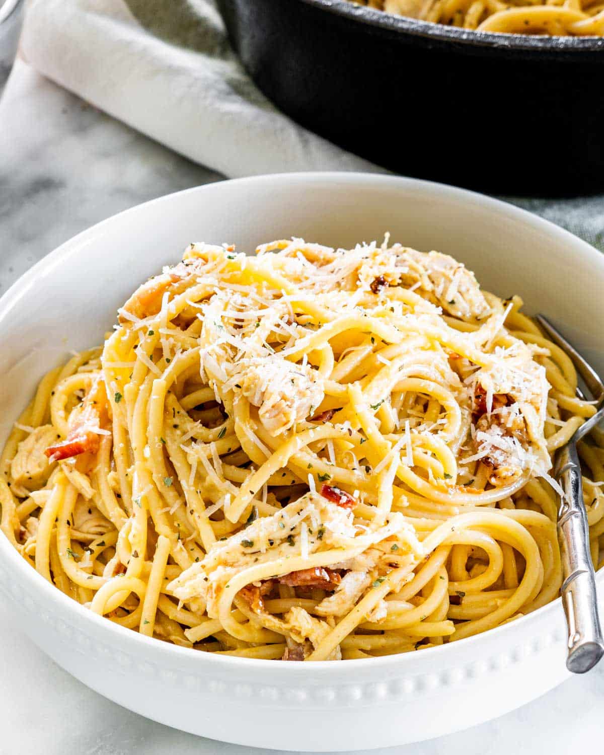 sideview shot of chicken carbonara in a white bowl with a fork in it, garnished with parmesan cheese