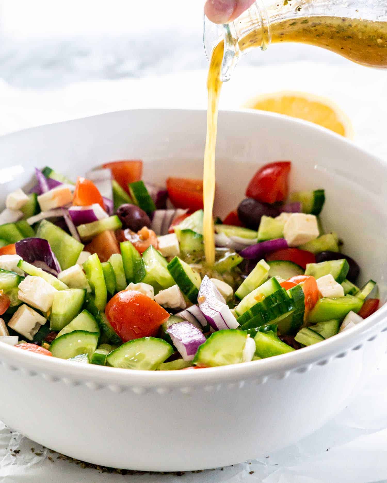 pouring of greek salad dressing onto a greek salad in a white bowl.