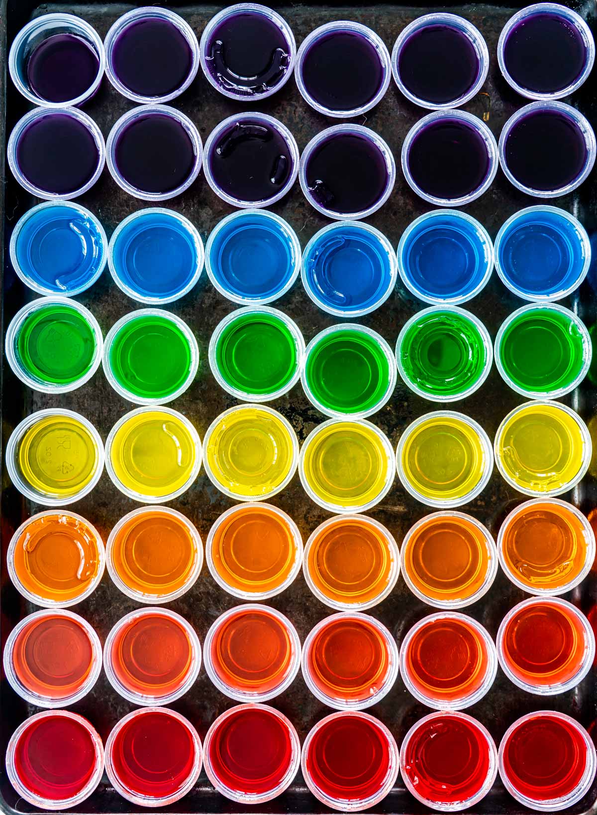 overhead shot of a rainbow of jello shots in little plastic cups with lids off