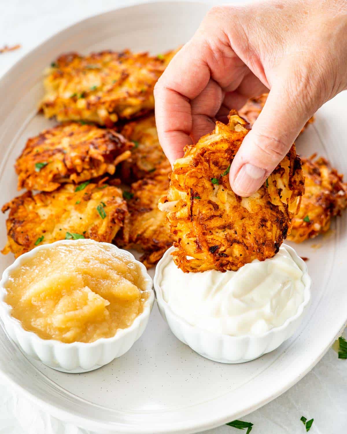 a hand dipping a latke in sour cream on a platter full of latkes