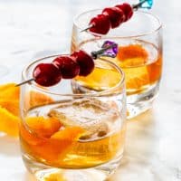 side view shot of two glasses of an old fashioned cocktail garnished with bourbon cherries on a pick and orange peel