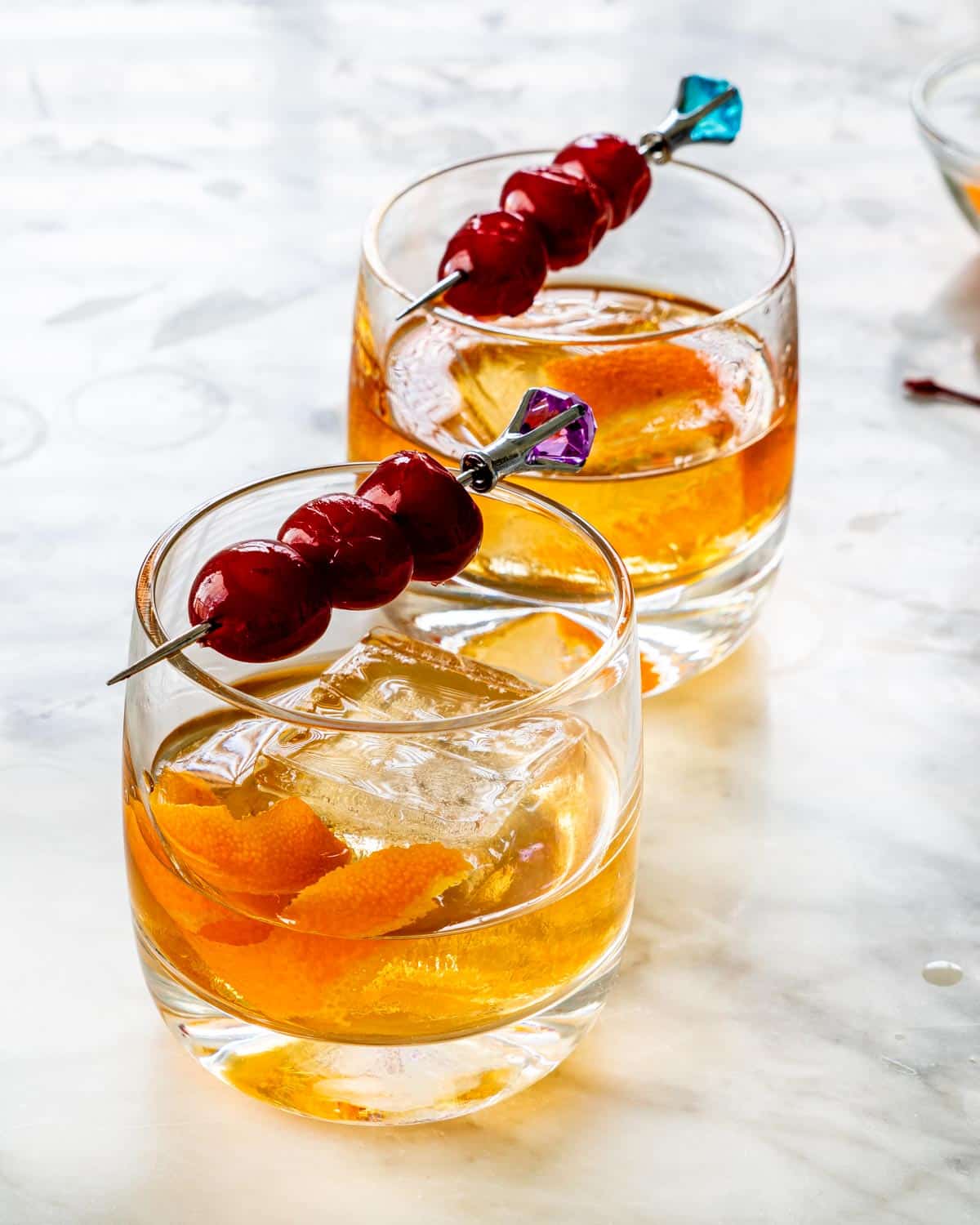 side view shot of two glasses of an old fashioned cocktail garnished with bourbon cherries on a pick and orange peel