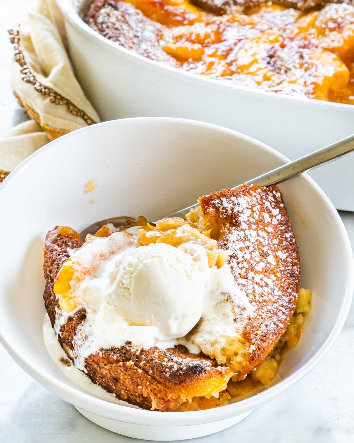 sideview shot of peach cobbler in a white bowl with a scoop of ice cream and a spoon inside