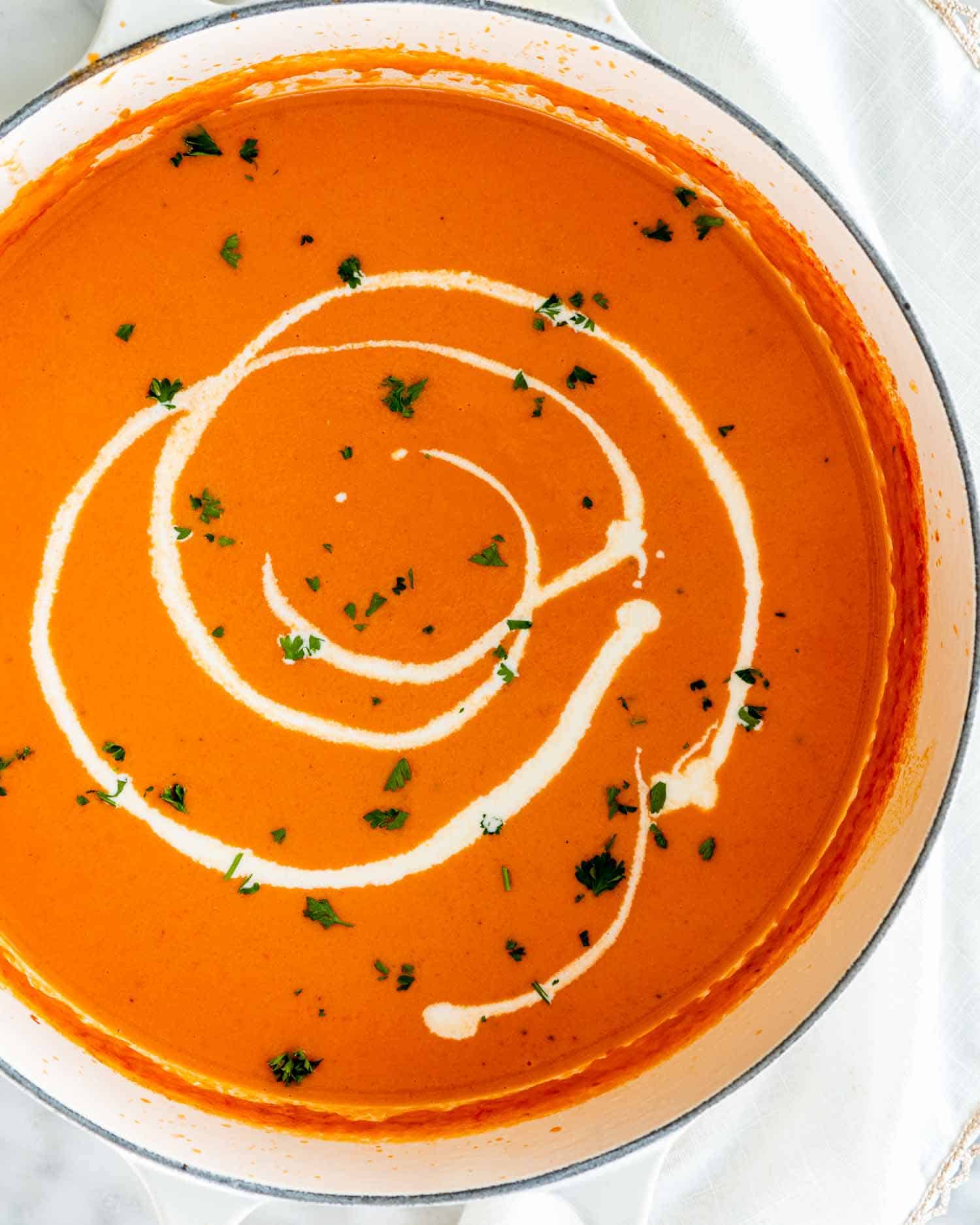 overhead shot of tomato bisque soup in a beige dutch oven with a drizzle of heavy cream garnished with parsley
