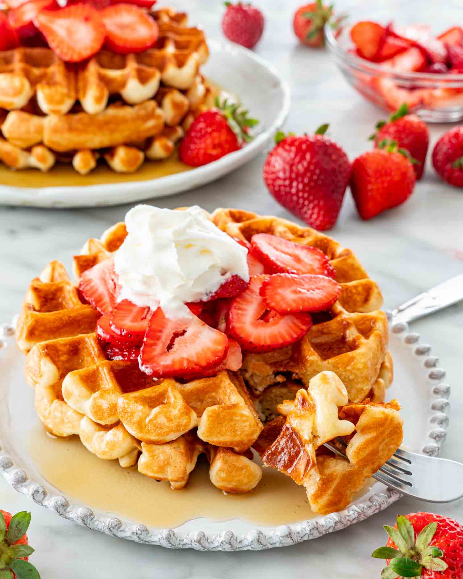two belgian waffles on a white plate topped with maple syrup, strawberries and whipped cream and a fork holding a piece.