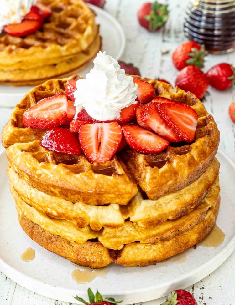 a stack of belgian waffles on a white plate topped with maple syrup, strawberries and whipped cream