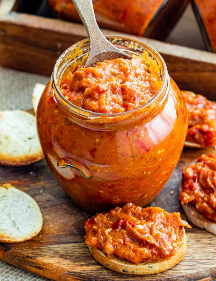 roasted eggplant and pepper spread in a jar with a wooden spoon inside surrounded by sliced of bread with zacusca