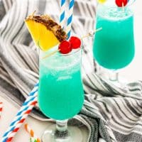 two glasses with blue hawaiian garnished with pineapple slice and maraschino cherries.