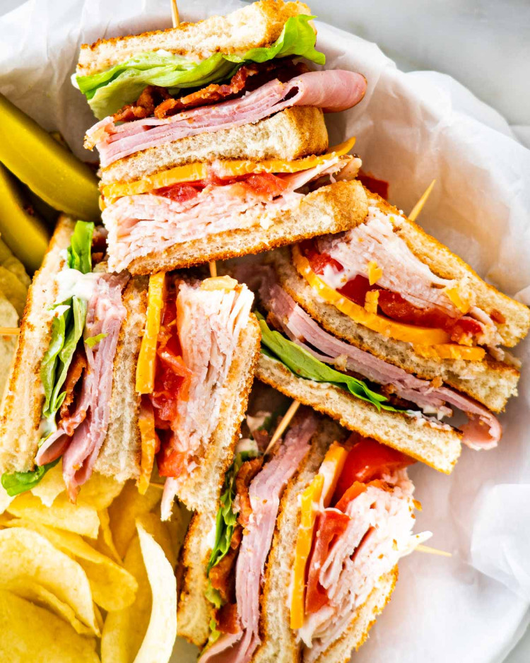 overhead of a club sandwich cut in quarters with a side of chips and pickles in a basket.