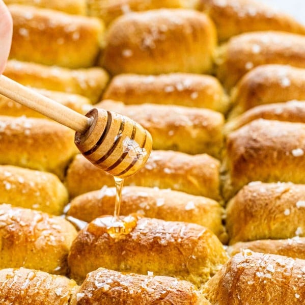 a hand drizzling honey over a pan full of parker house rolls.