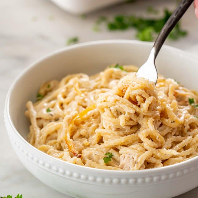 A fork lifts creamy spaghetti from turkey tetrazzini in a white beaded bowl, with parsley scattered on the marble surface.