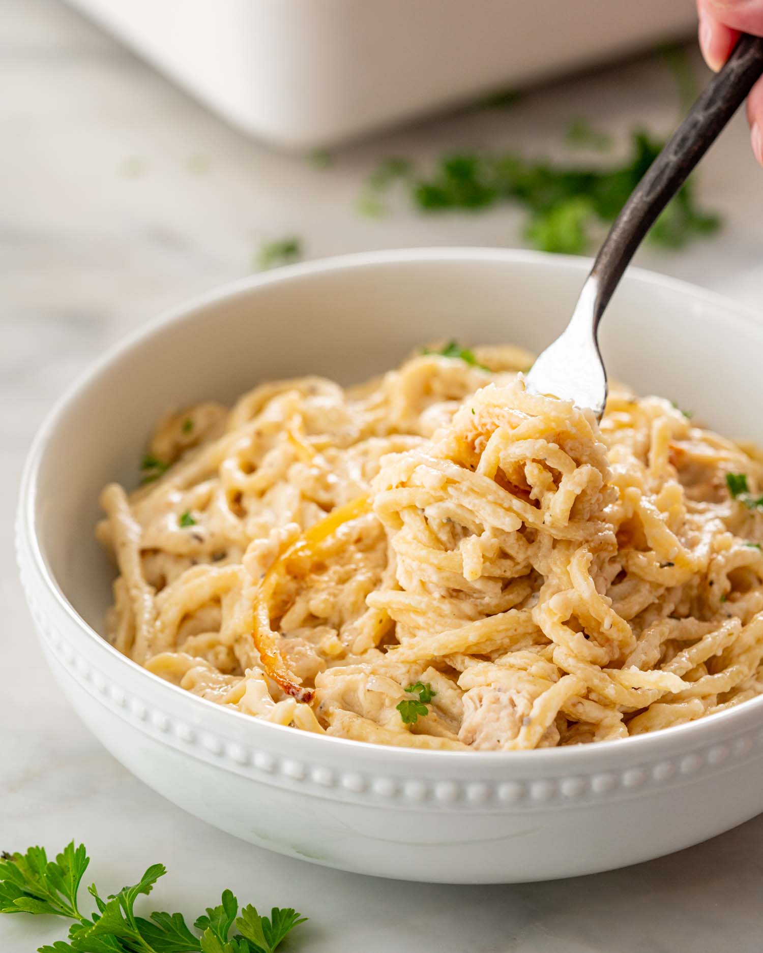 A fork lifts creamy spaghetti from turkey tetrazzini in a white beaded bowl, with parsley scattered on the marble surface.