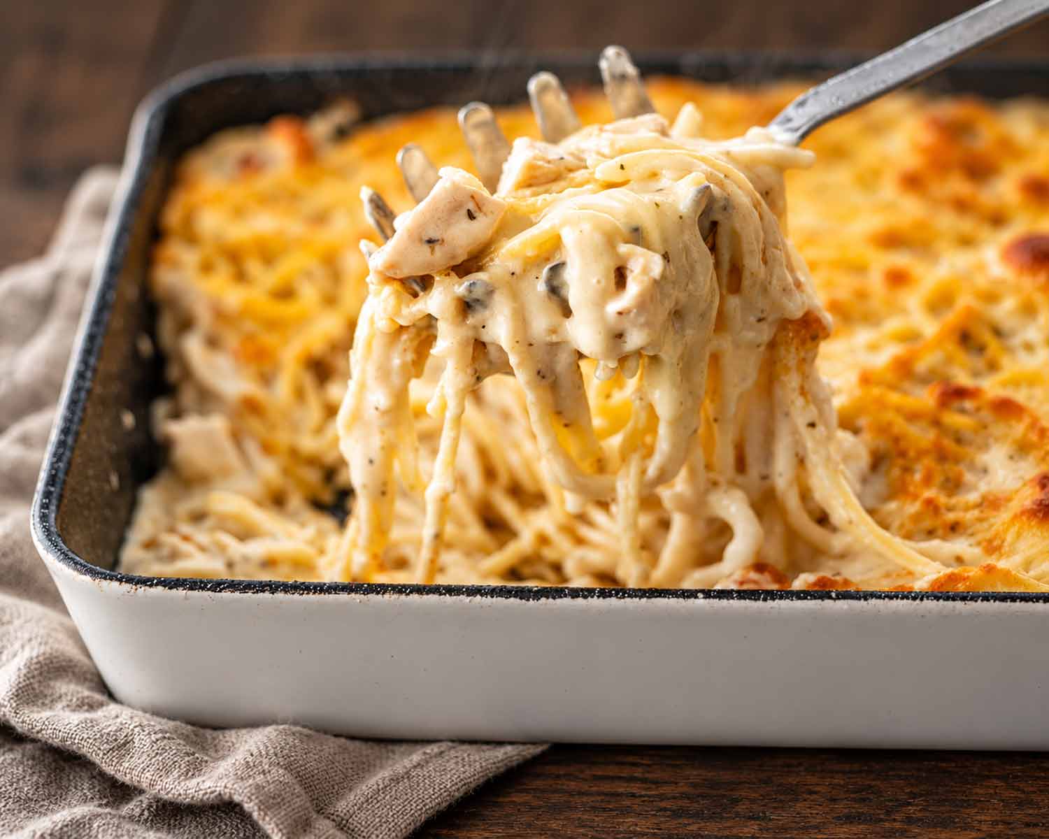 Close-up of creamy turkey tetrazzini lifted on a pasta server above a golden casserole, with walnut wood and oatmeal linen.