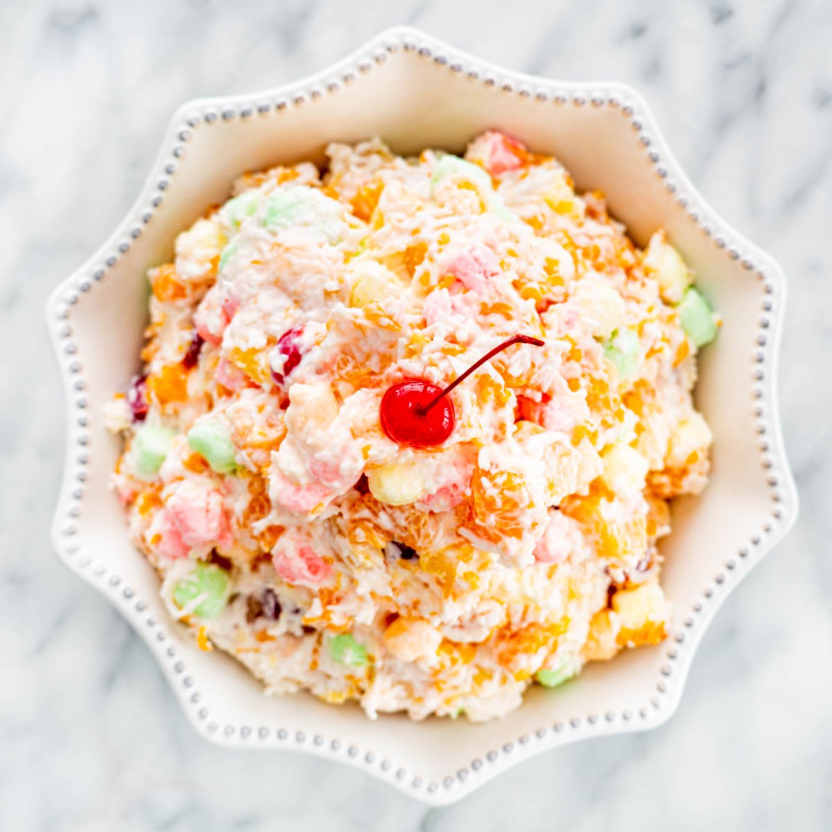 overhead of a a white bowl piled high with ambrosia salad and a maraschino cherry on top.