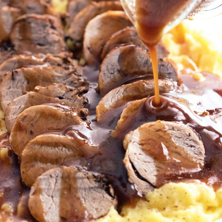gravy being poured over sliced pork loin on top of a bed of mashed potatoes