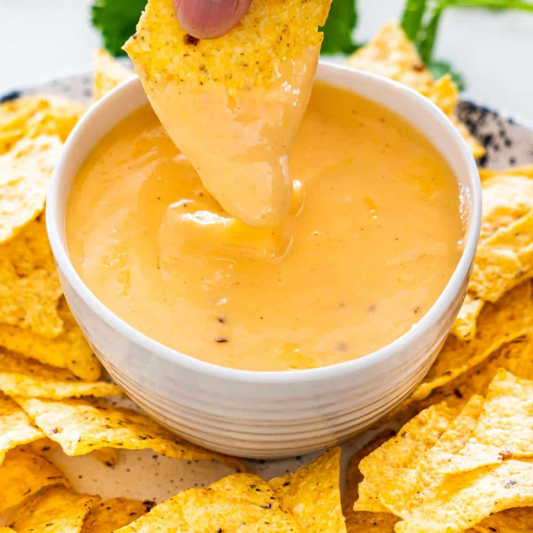 a hand dipping a tortilla chip in a bowl of nacho cheese sauce.