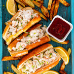 3 lobster rolls on a blue tray with french fries around and garnished with lemon wedges.