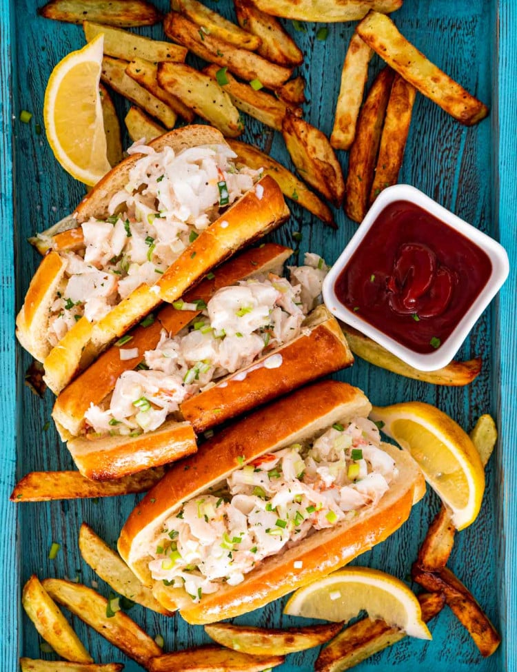3 lobster rolls on a blue tray with french fries around and garnished with lemon wedges.