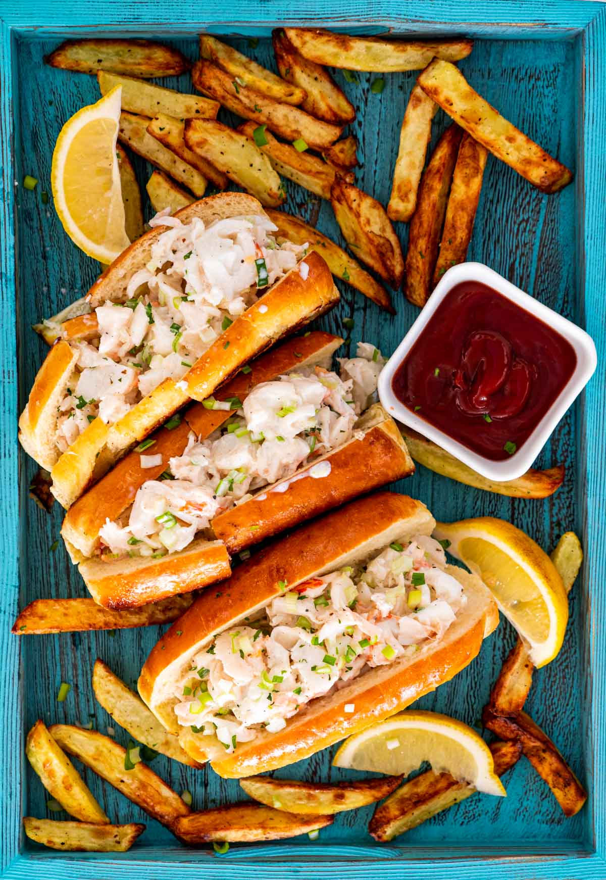 3 lobster rolls on a blue tray with french fries around and garnished with lemon wedges.