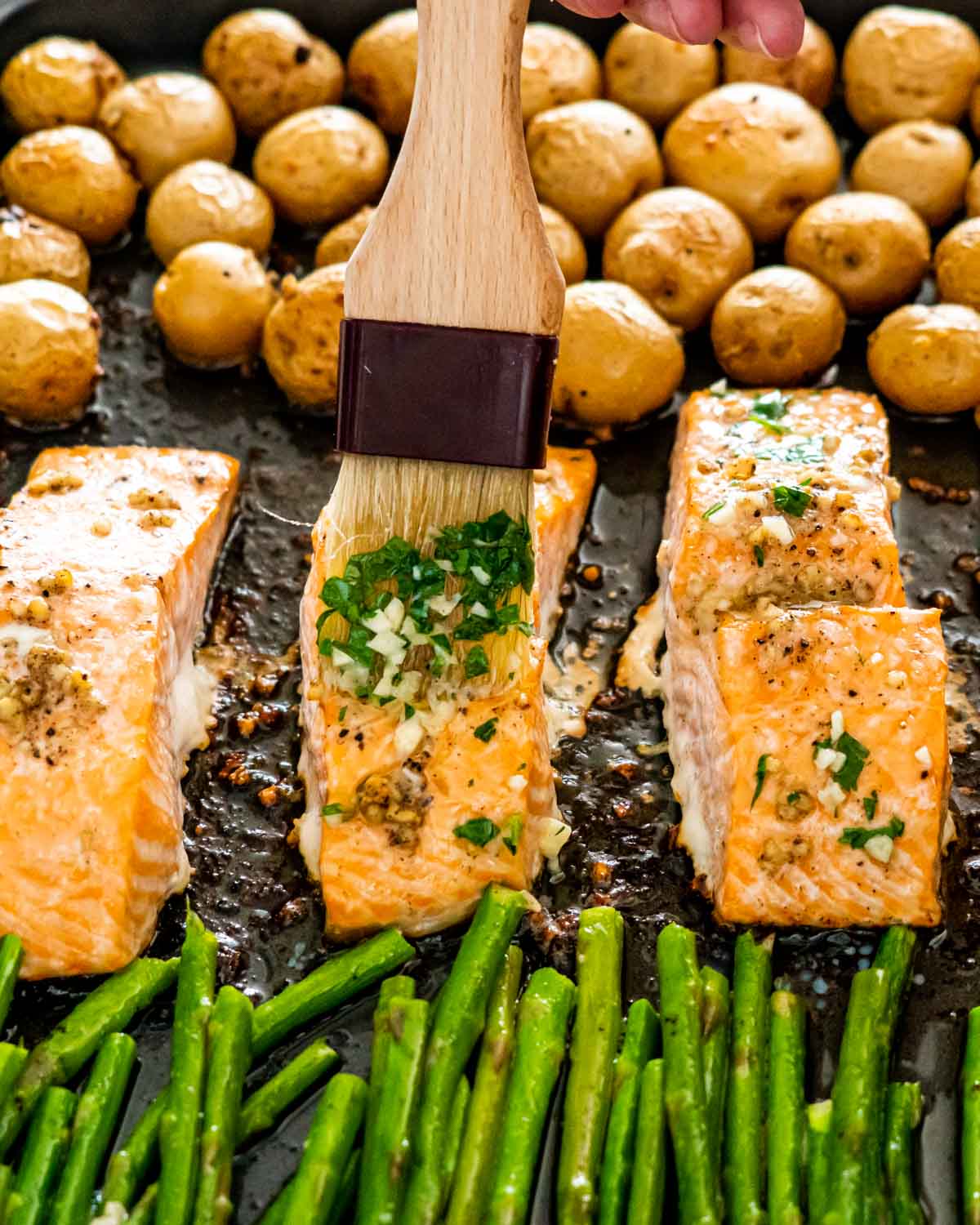 a hand holding a pastry brush and brushing garlic butter sauce over salmon on a sheet pan.