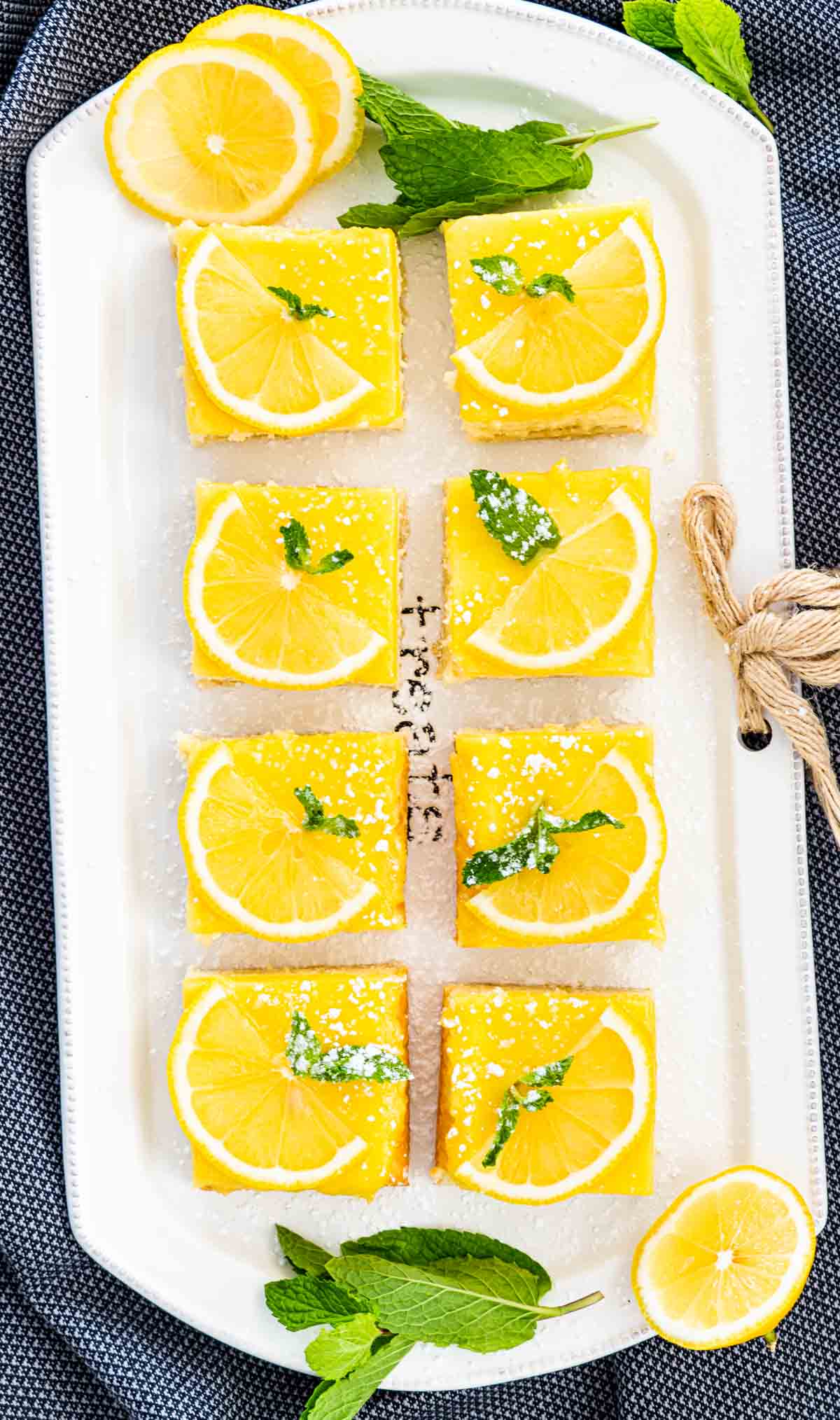 a bunch of lemon cheesecake bars on a white serving platter garnished with lemon slices and mint leaves.