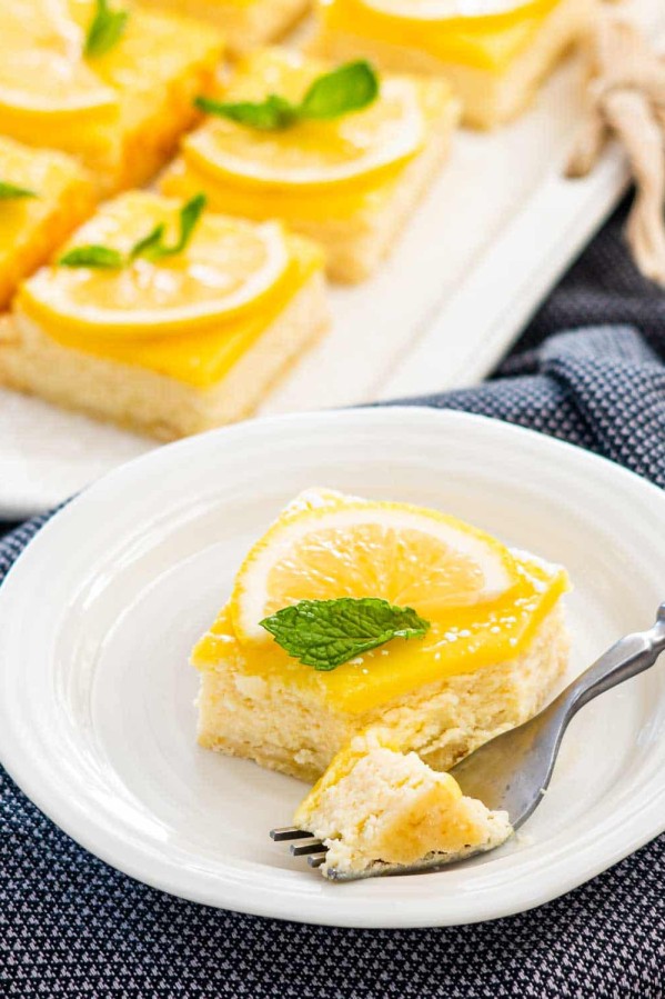 a lemon cheesecake bar garnished with lemon slice and mint leaf on a white plate with a fork taking out a piece.