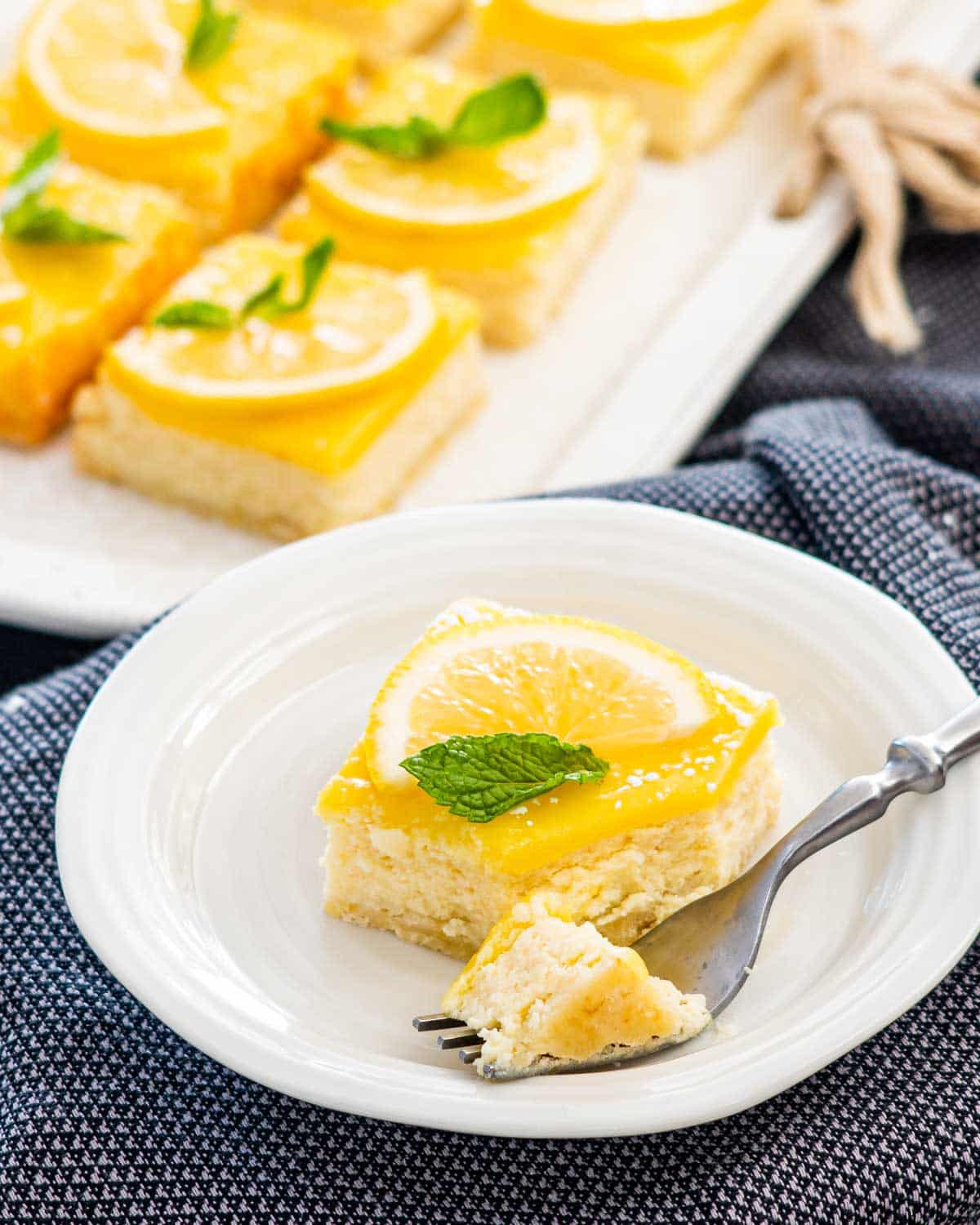 a lemon cheesecake bar garnished with lemon slice and mint leaf on a white plate with a fork taking out a piece.