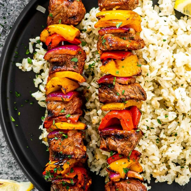 2 skewers of beef shish kebabs on a plate next to a bed of rice on a black plate.