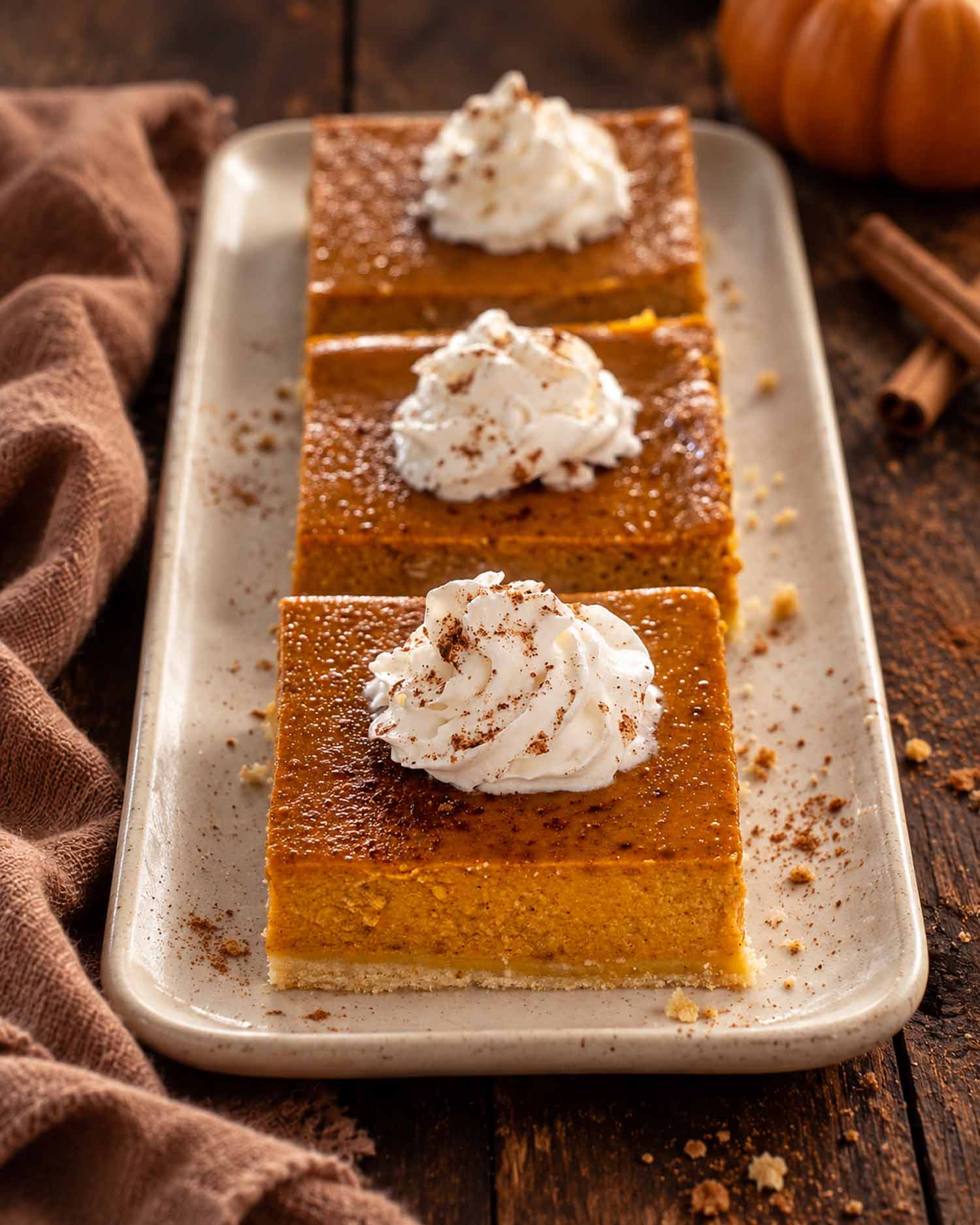 Three pumpkin pie bars topped with whipped cream and cinnamon on a cream platter beside linen and cinnamon sticks.