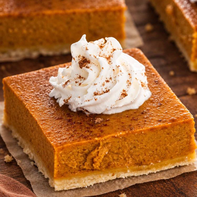 Close-up of a pumpkin pie bar with shortbread crust and cinnamon-dusted whipped cream on parchment over a wooden table.