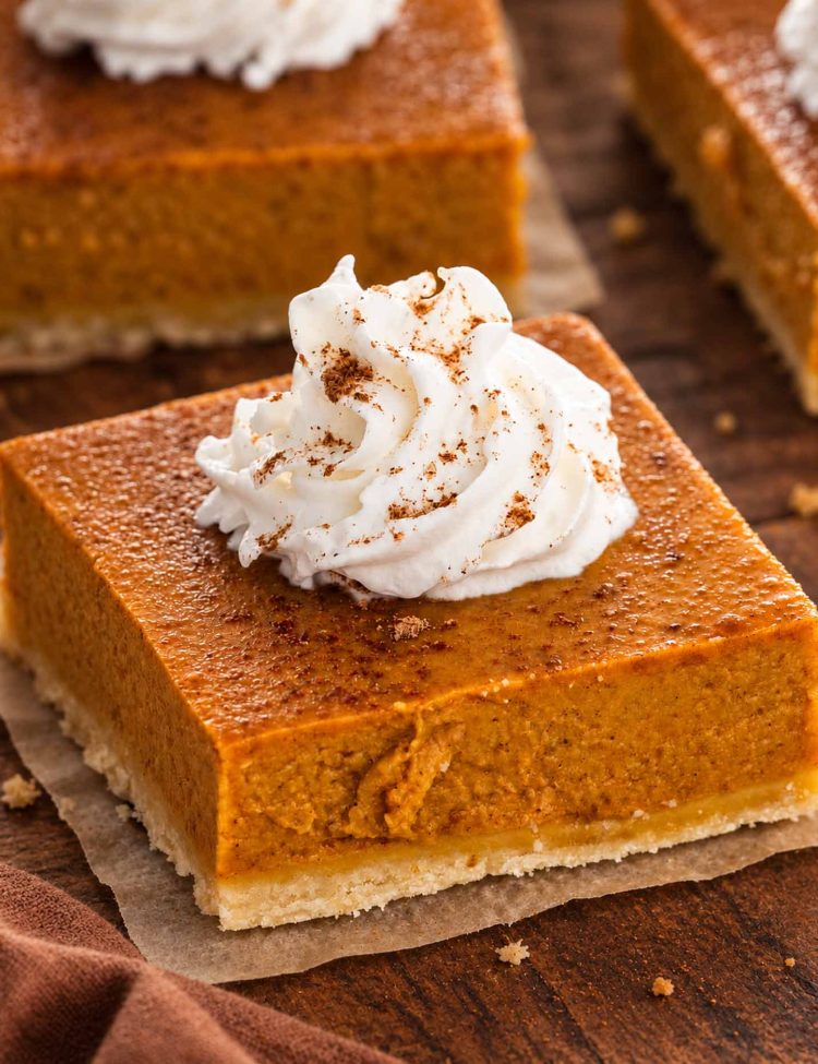 Close-up of a pumpkin pie bar with shortbread crust and cinnamon-dusted whipped cream on parchment over a wooden table.