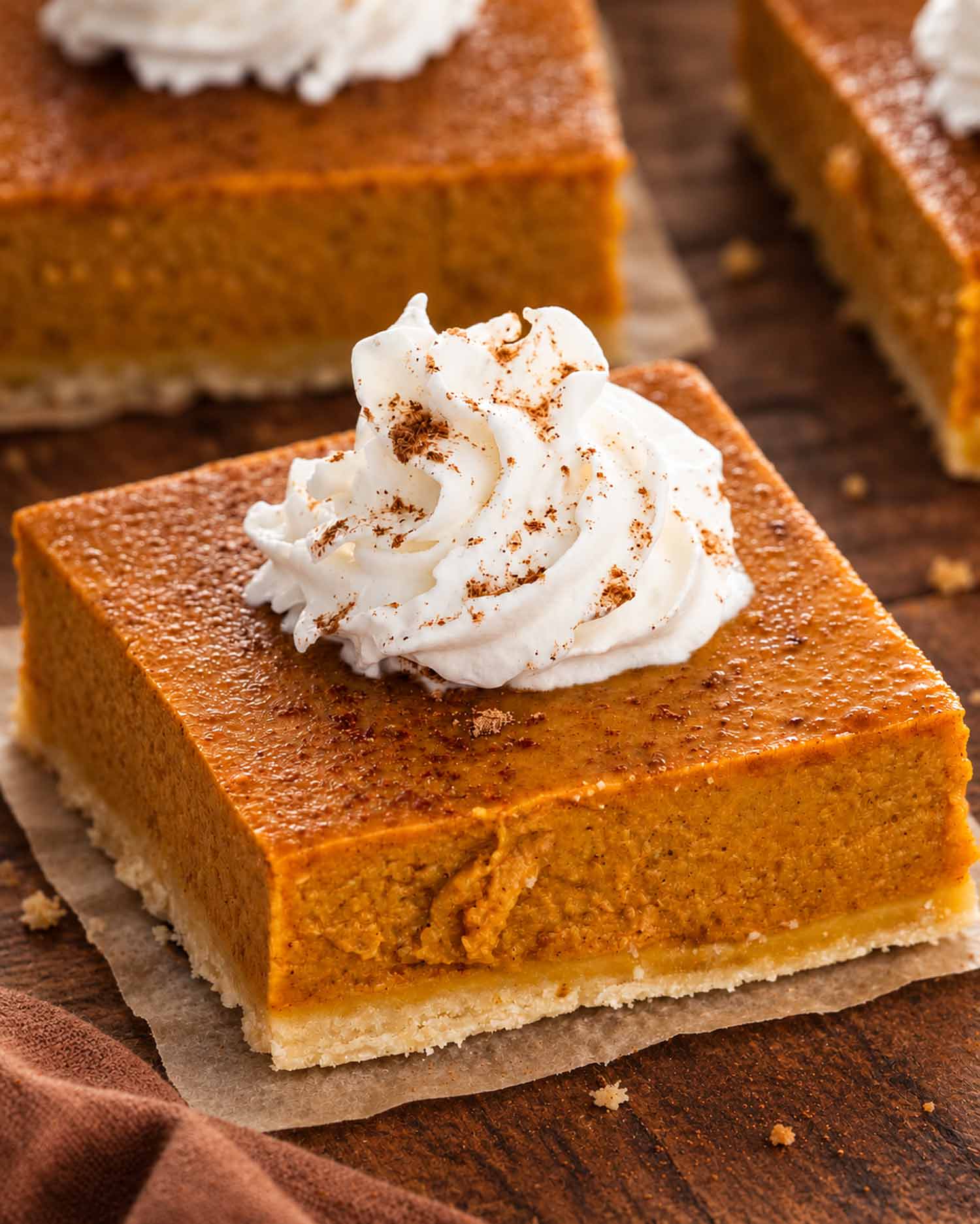 Close-up of a pumpkin pie bar with shortbread crust and cinnamon-dusted whipped cream on parchment over a wooden table.
