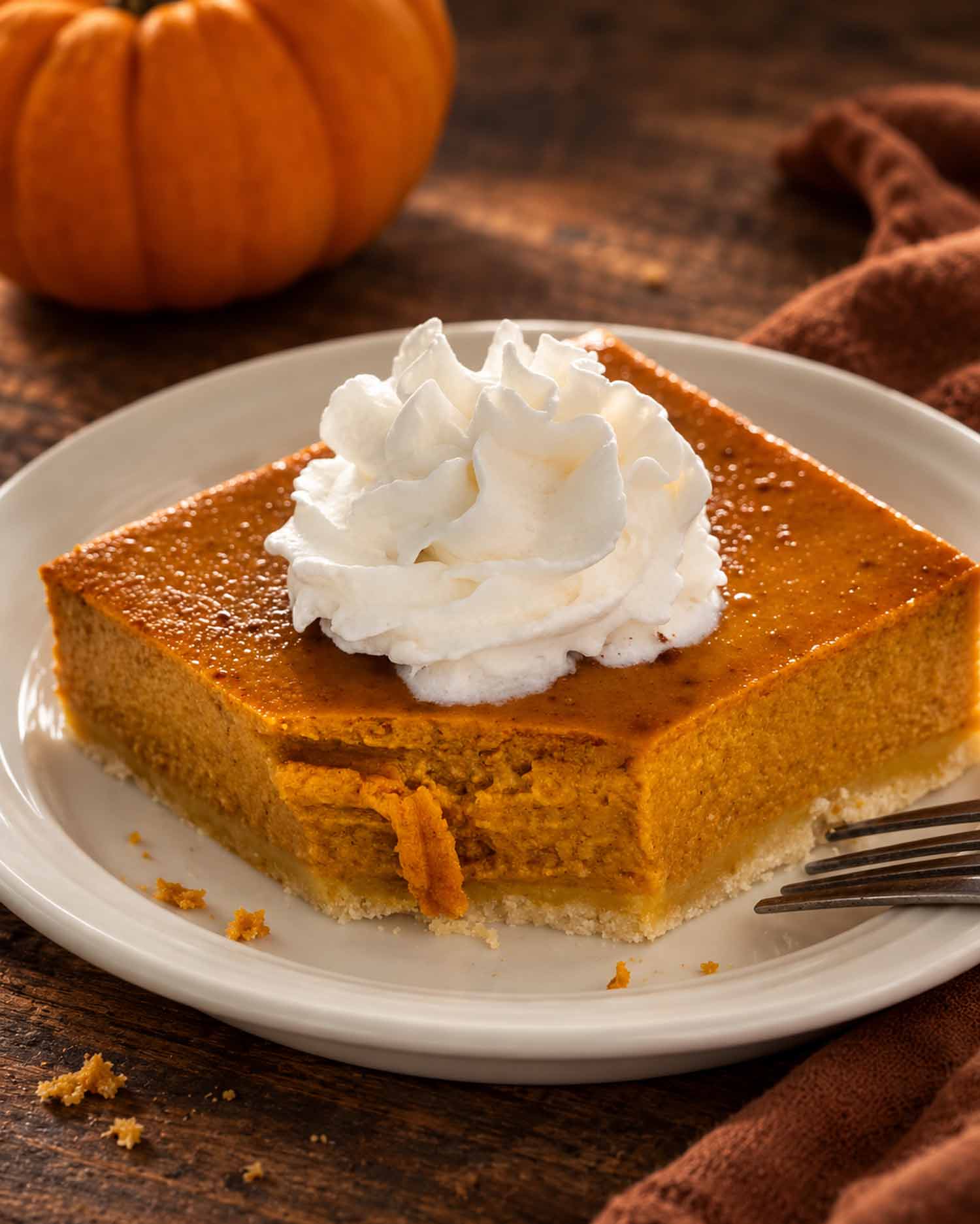 Pumpkin pie bar topped with whipped cream on a white plate with a fork, pumpkin, and rust-colored linen nearby.