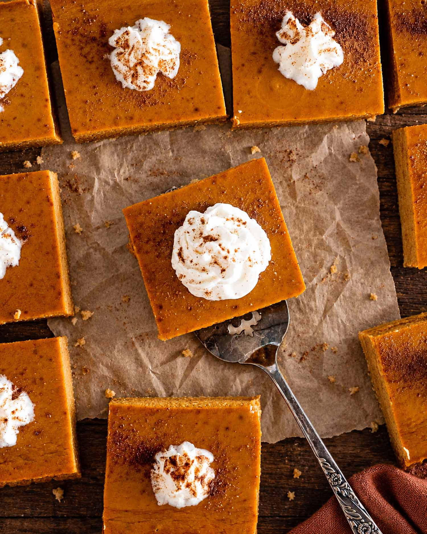 Overhead view of pumpkin pie bars topped with cinnamon-dusted whipped cream on parchment, with a vintage server beneath one square.