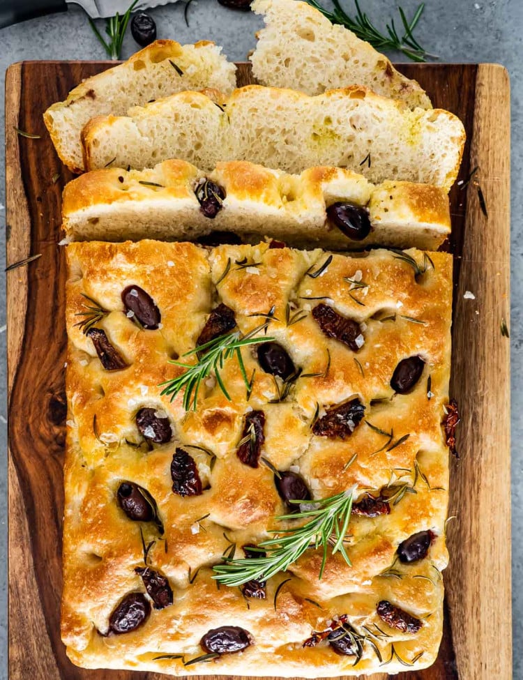 focaccia with sun dried tomatoes and olives on a cutting board with a couple slices cut out.