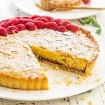 a bakewell tart on a serving platter with a slice missing garnished with raspberries.