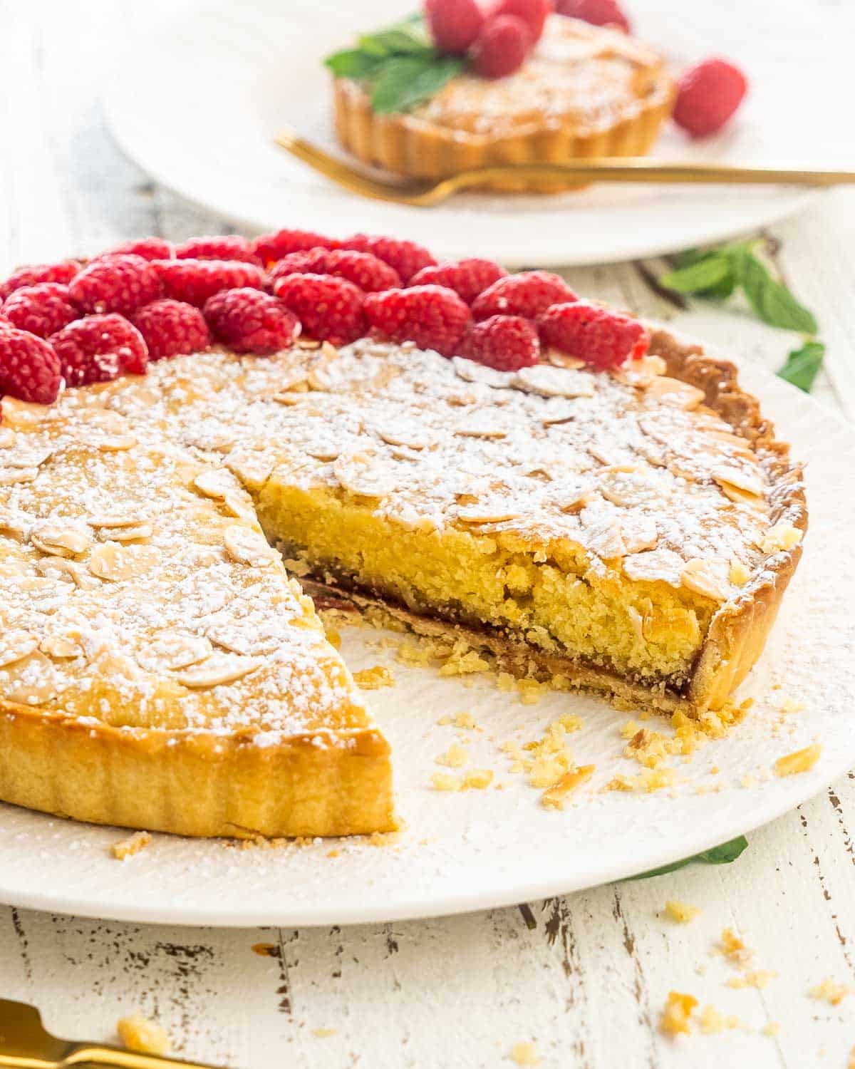 a bakewell tart on a serving platter with a slice missing garnished with raspberries.