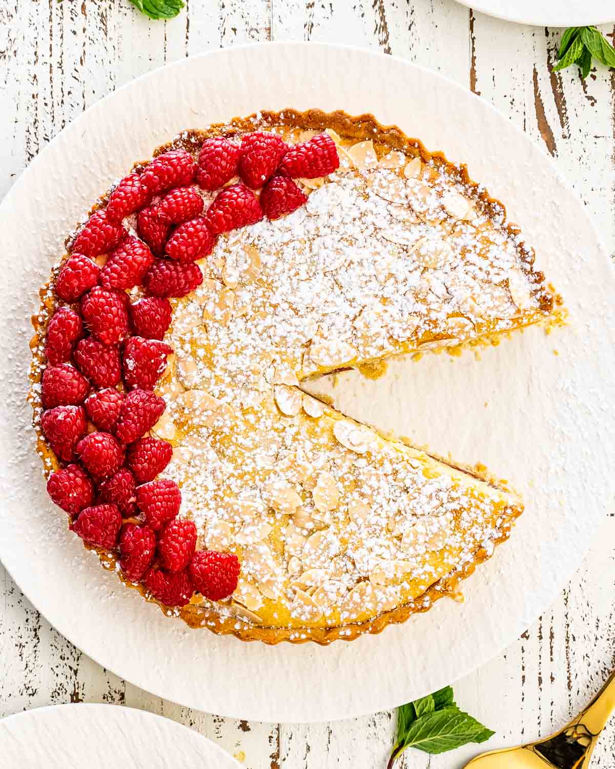 a bakewell tart on a serving platter with a slice missing garnished with raspberries.