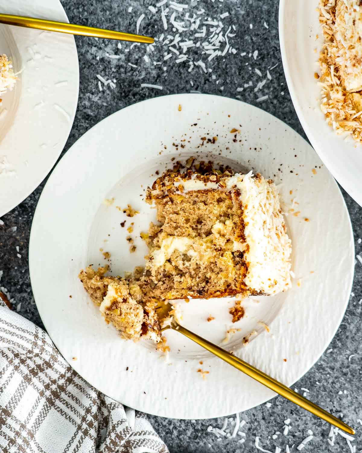 a slice of hummingbird cake on a white plate with a gold fork taking a bite out of it.