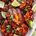 grilled pollo asado on a white platter garnished with cilantro and lime wedges.