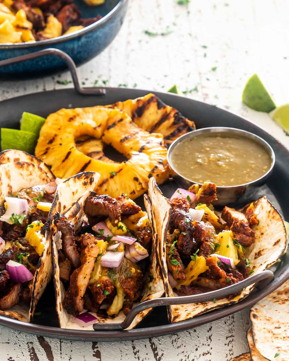 tacos al pastor with grilled pineapple slices and green salsa on a serving platter.