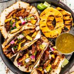 tacos al pastor with grilled pineapple slices and green salsa on a serving platter.