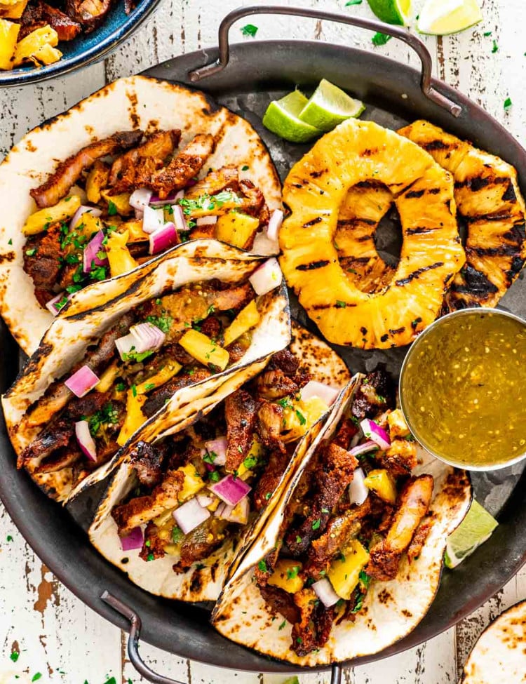 tacos al pastor with grilled pineapple slices and green salsa on a serving platter.