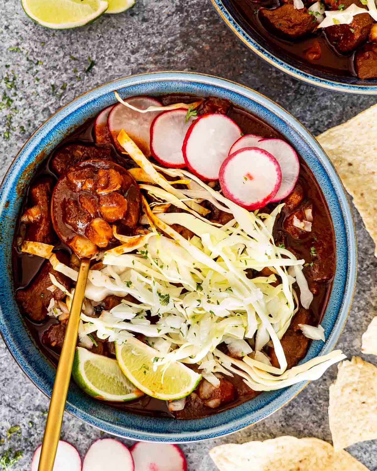 pozole rojo in a blue bowl with cabbage, radishes, onion and lime wedges.