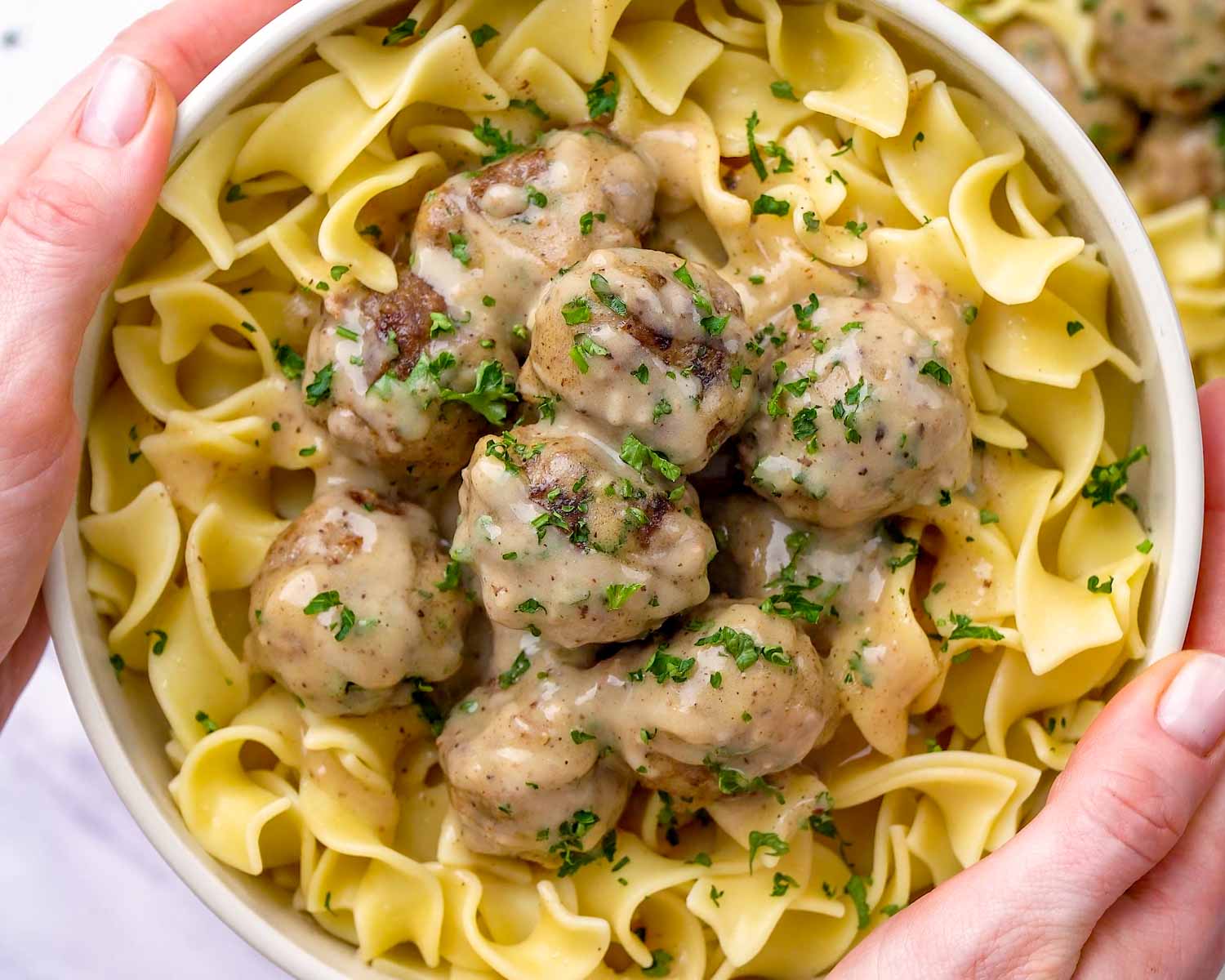 Hands holding a white bowl filled with wide egg noodles topped with eight beef Swedish meatballs and creamy gravy with fresh parsley.