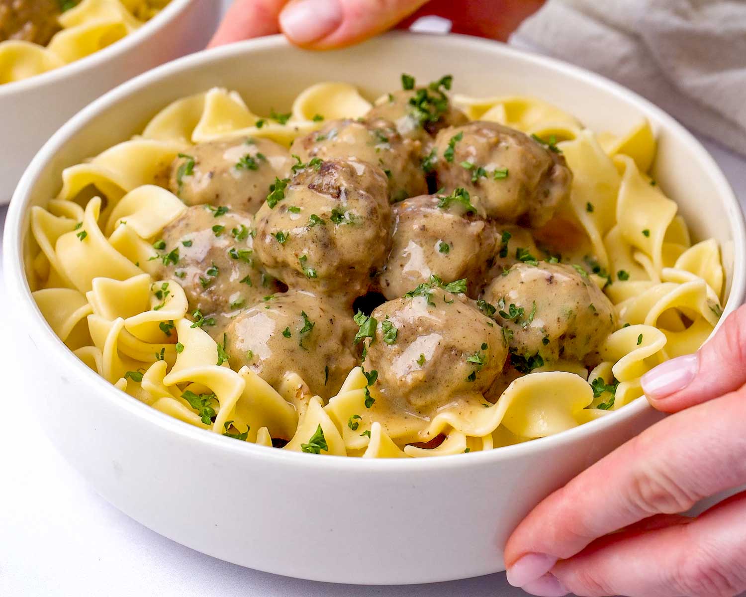 Hands holding a white bowl filled with wide egg noodles topped with eight beef Swedish meatballs and creamy gravy with fresh parsley.
