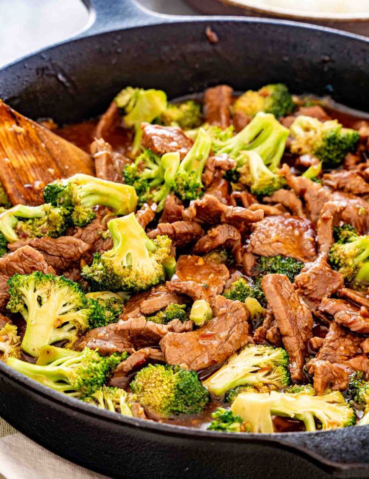 Beef strips and green broccoli florets glistening with sauce in a black skillet, with a wooden spatula tucked alongside.