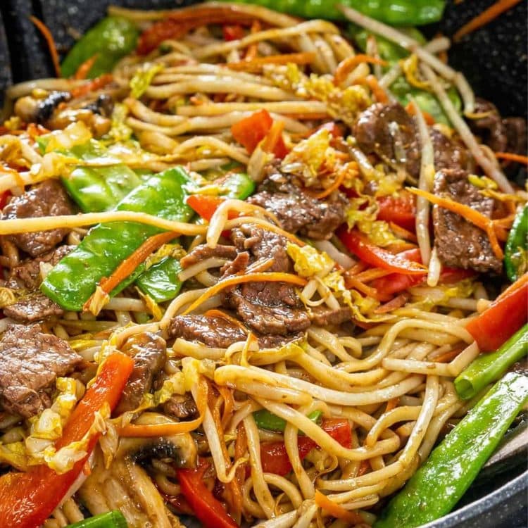 Close up of saucy beef lo mein noodles with tender steak strips, snow peas, and carrots in a dark wok.