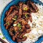crispy sticky beef with rice garnished with green onions and sesame seeds in a blue bowl.
