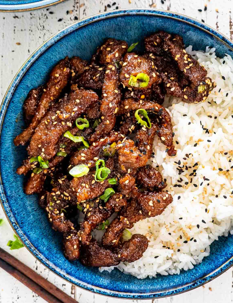 crispy sticky beef with rice garnished with green onions and sesame seeds in a blue bowl.